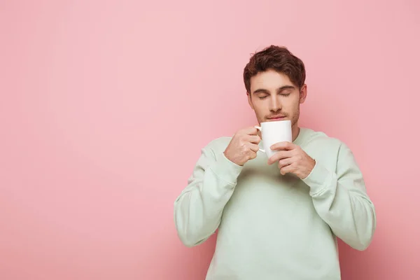 Красивый молодой человек наслаждается ароматом кофе, держа белую чашку на розовом фоне — стоковое фото