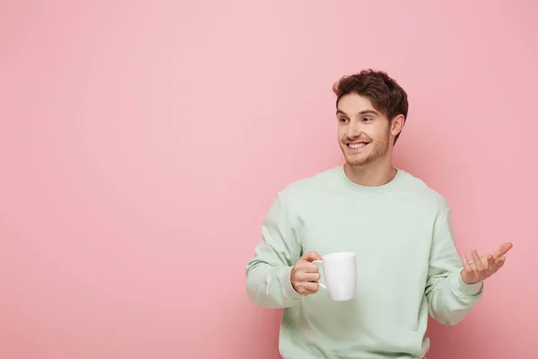 Alegre joven sosteniendo la taza mientras está de pie con el brazo abierto y mirando hacia otro lado sobre fondo rosa - foto de stock