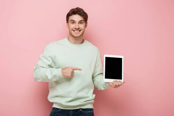 Sonriente hombre apuntando con el dedo a la tableta digital con pantalla en blanco mientras mira a la cámara en el fondo rosa - foto de stock