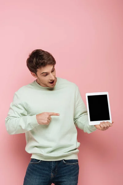 Hombre sorprendido apuntando con el dedo a la tableta digital con pantalla en blanco sobre fondo rosa - foto de stock