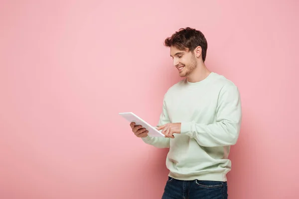 Schöner Kerl lächelt, während er digitales Tablet auf rosa Hintergrund benutzt — Stockfoto