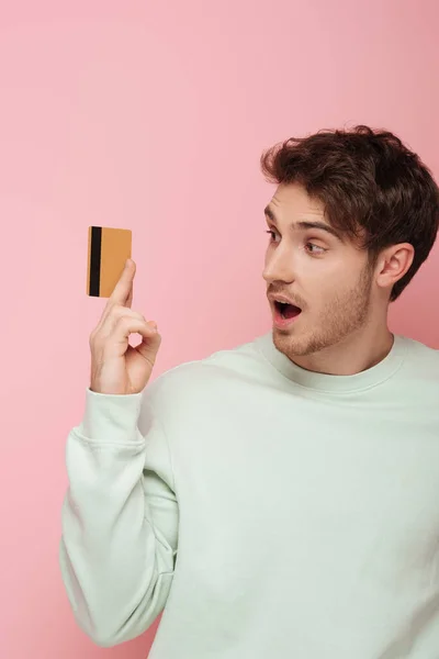 Surprised young man looking at credit card on pink background — Stock Photo