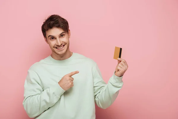 Sonriente joven apuntando con el dedo a la tarjeta de crédito mientras mira a la cámara en el fondo rosa - foto de stock