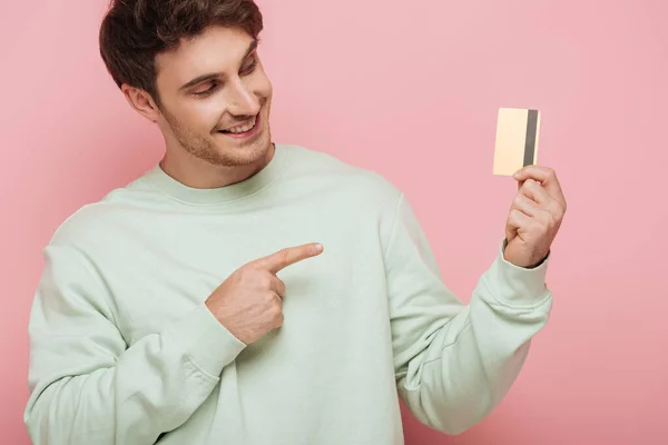 Sonriente joven apuntando con el dedo a la tarjeta de crédito sobre fondo rosa - foto de stock