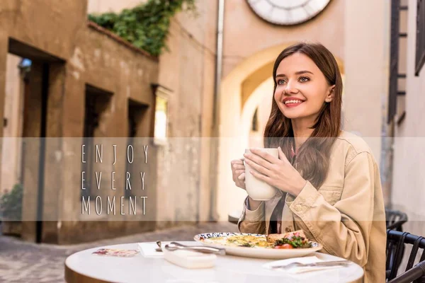 Selective focus of woman smiling and holding white cup of tea in cafe in city, enjoy every moment illustration — Stock Photo