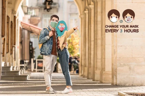 Couple of tourists with illustrated faces in medical masks hugging on street and pointing with finger away, change your mask every 3 hours illustration — Stock Photo