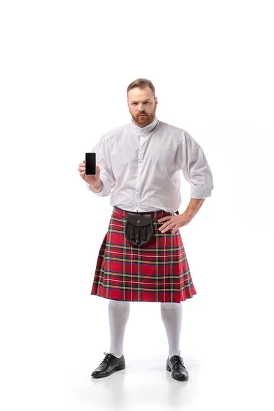 Grave escocês ruiva homem em vermelho kilt segurando smartphone com tela em branco no fundo branco — Fotografia de Stock