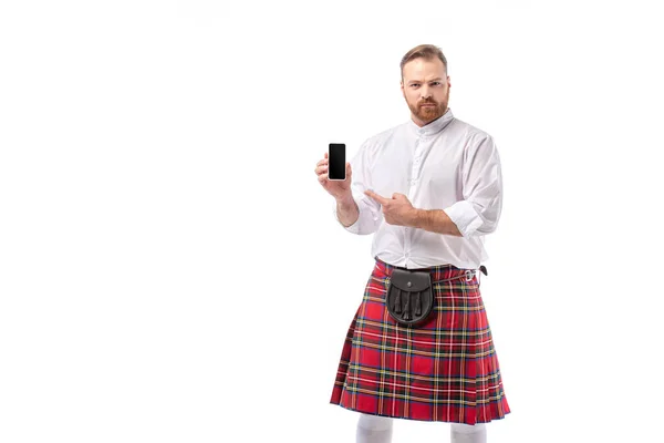 Serious Scottish redhead man in red kilt pointing at smartphone with blank screen isolated on white — Stock Photo