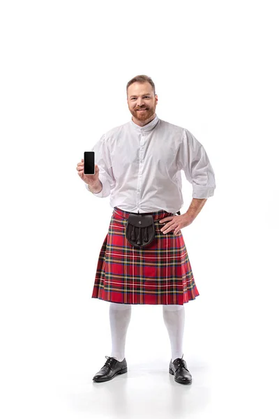 Улыбающийся шотландский рыжий мужчина в красном килте держит смартфон с чистым экраном на белом фоне — стоковое фото