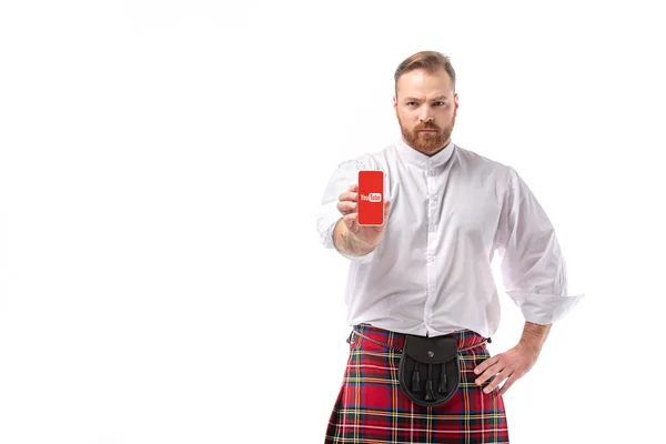 KYIV, UKRAINE - NOVEMBER 29, 2019: serious Scottish redhead man in red kilt presenting smartphone with youtube app isolated on white — Stock Photo