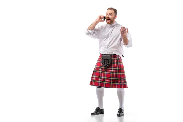 Animado escocês ruiva homem em kilt vermelho falando no smartphone no fundo branco — Fotografia de Stock
