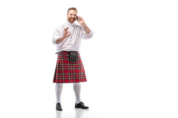 Happy Scottish redhead man in red kilt talking on smartphone and showing ok sign on white background — Stock Photo