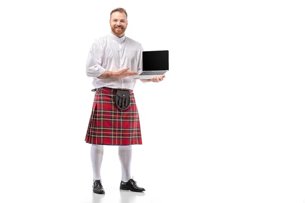 Souriant écossais rousse homme en kilt rouge tenant ordinateur portable avec écran blanc sur fond blanc — Photo de stock