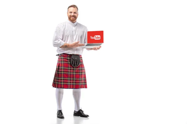 KYIV, UKRAINE - 29 NOVEMBRE 2019 : homme rousse écossais souriant en kilt rouge présentant un ordinateur portable avec le site youtube sur fond blanc — Photo de stock