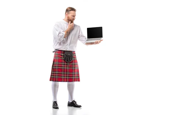 Pensive Scottish redhead man in red kilt holding laptop with blank screen on white background — Stock Photo