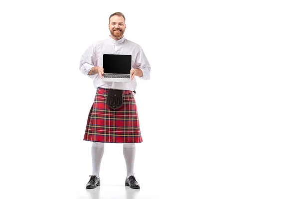 Sonriente escocés pelirroja hombre en escocés rojo presentando portátil con pantalla en blanco sobre fondo blanco - foto de stock