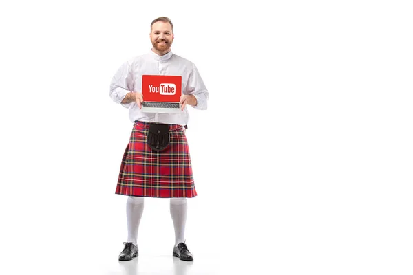 KYIV, UCRANIA - 29 de noviembre de 2019: pelirrojo escocés sonriente en escocés rojo presentando portátil con sitio web de youtube sobre fondo blanco - foto de stock