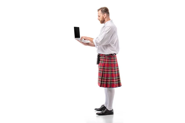 Vue latérale de l'homme rousse écossais en kilt rouge à l'aide d'un ordinateur portable sur fond blanc — Photo de stock