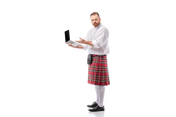 Grave escocês ruiva homem no vermelho kilt segurando laptop no fundo branco — Fotografia de Stock