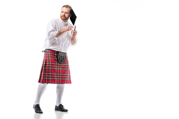 Hombre pelirrojo escocés en escocés rojo sosteniendo portátil cerca de la oreja sobre fondo blanco - foto de stock