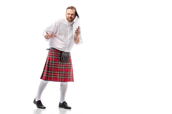 Confuso hombre pelirrojo escocés en escocés rojo sosteniendo portátil cerca de la oreja sobre fondo blanco - foto de stock