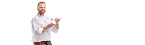 Smiling Scottish redhead man in red kilt drinking coffee isolated on white, panoramic crop — Stock Photo