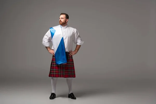 Serious Scottish redhead man in red kilt with flag of Scotland and hands on hips on grey background — Stock Photo