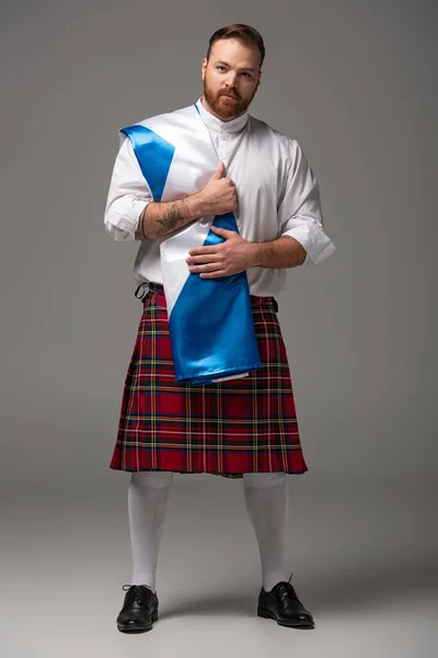 Scottish redhead man in red kilt with flag of Scotland on grey background — Stock Photo