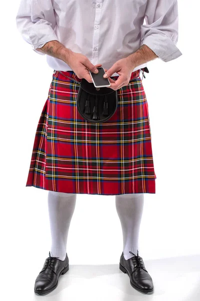 Обрезанный вид шотландца в красном кильте с кожаной сумкой и смартфоном на белом фоне — стоковое фото