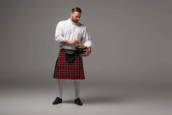 Scottish redhead man in red kilt holding potty with gold coins on grey background — Stock Photo