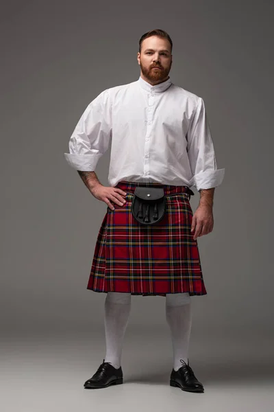 Scozzese rossa uomo in kilt rosso con mano sul fianco su sfondo grigio — Foto stock