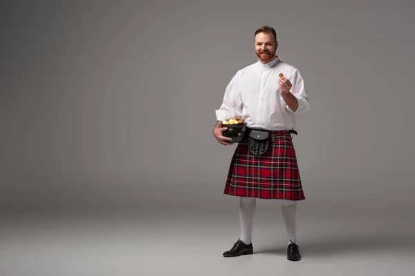 Sorridente scozzese rossa uomo in kilt rosso che tiene vasino con monete d'oro su sfondo grigio — Foto stock