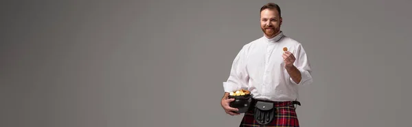 Lächelnder schottischer Rotschopf im roten Kilt mit Töpfchen mit Goldmünzen auf grauem Hintergrund, panoramische Ernte — Stockfoto