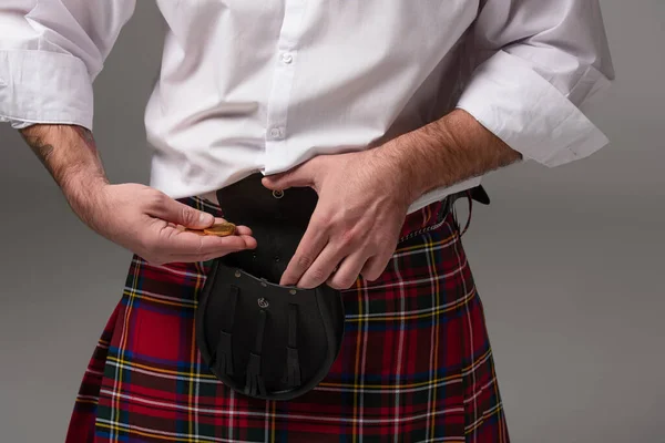 Partial view of Scottish man in red kilt putting gold coins in belt bag isolated on grey — Stock Photo