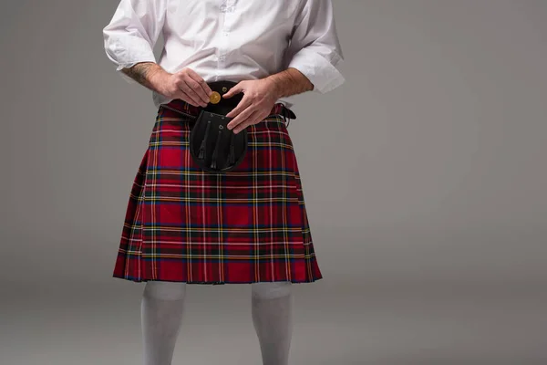Vue recadrée de l'homme rousse écossais en kilt rouge mettant des pièces d'or dans un sac de ceinture sur fond gris — Photo de stock
