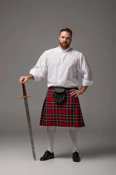 Scottish redhead man in red kilt with sword on grey background — Stock Photo