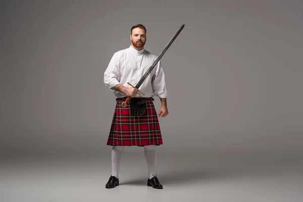 Шотландский рыжий человек в красном килте с мечом на сером фоне — стоковое фото
