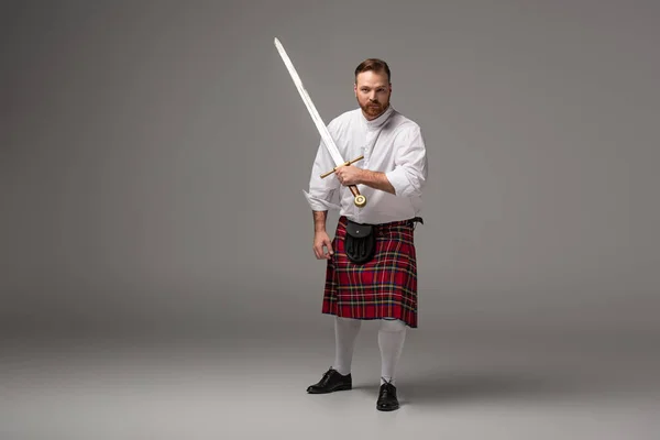 Escocês ruiva homem em kilt vermelho com espada no fundo cinza — Fotografia de Stock