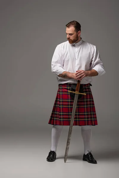 Escocês ruiva homem em kilt vermelho com espada no fundo cinza — Fotografia de Stock