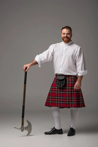 Sorridente scozzese rossa uomo in kilt rosso con ascia da battaglia su sfondo grigio — Foto stock