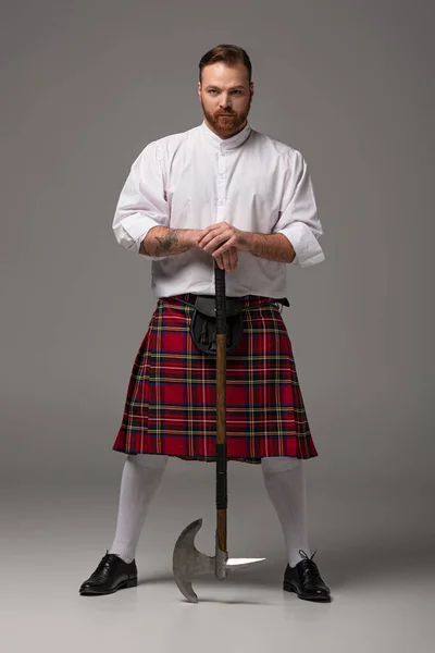 Serious Scottish redhead man in red kilt with battle axe on grey background — Stock Photo