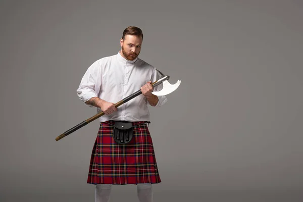 Serious Scottish redhead man in red kilt with battle axe on grey background — Stock Photo