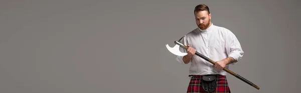 Serious Scottish redhead man in red kilt with battle axe on grey background, panoramic shot — Stock Photo