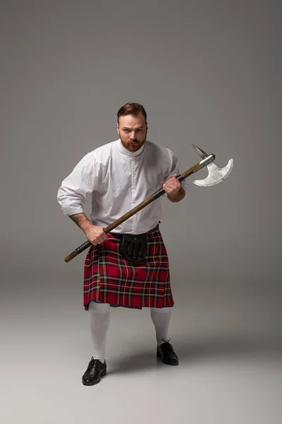 Сердитый шотландский рыжий мужчина в красном килте с боевым топором на сером фоне — стоковое фото