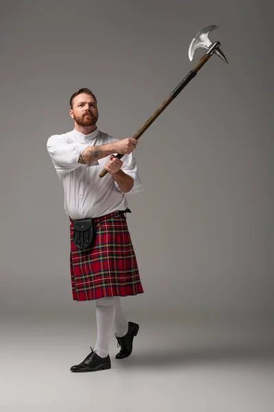 Escocês ruiva homem em kilt vermelho levantando machado de batalha no fundo cinza — Fotografia de Stock