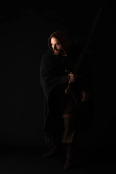 Medieval Scottish redhead man in mantel with sword on dark isolated on black — Stock Photo