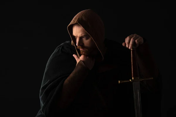 Frowning medieval Scottish man in mantel with sword  in dark isolated on black — Stock Photo