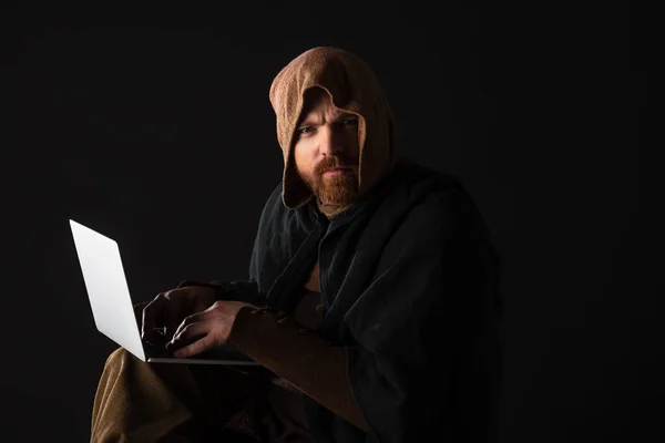 Frowning medieval Scottish man in mantel using laptop in dark isolated on black — Stock Photo