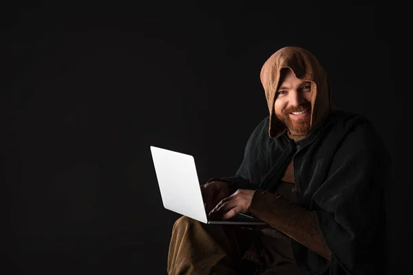 Souriant médiéval écossais en manteau en utilisant un ordinateur portable dans l'obscurité isolé sur noir — Photo de stock