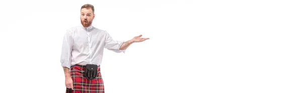 Удивленный шотландский рыжеволосый бородатый мужчина в красном тартановом килте указывая рукой, изолированной на белом, заголовок сайта — стоковое фото